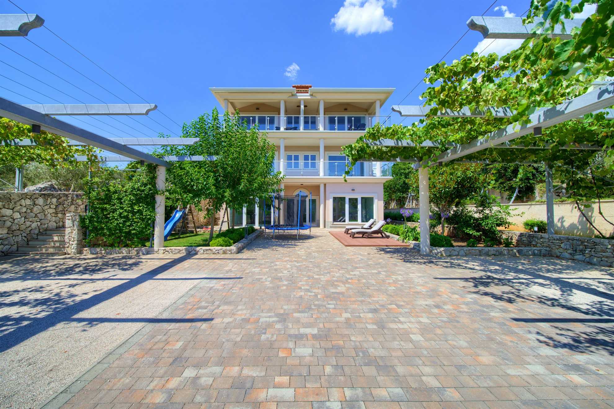 Apartment with a stone pathway surrounded by greenery. In front of the apartment are sun loungers, a slide and a trampoline