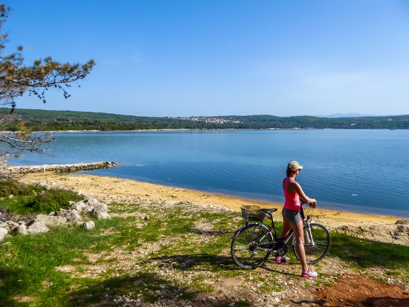 Djevojka s biciklom uživa na plažu gledajući more. U daljini je šumoviti predio u kojem se nadziru kućice