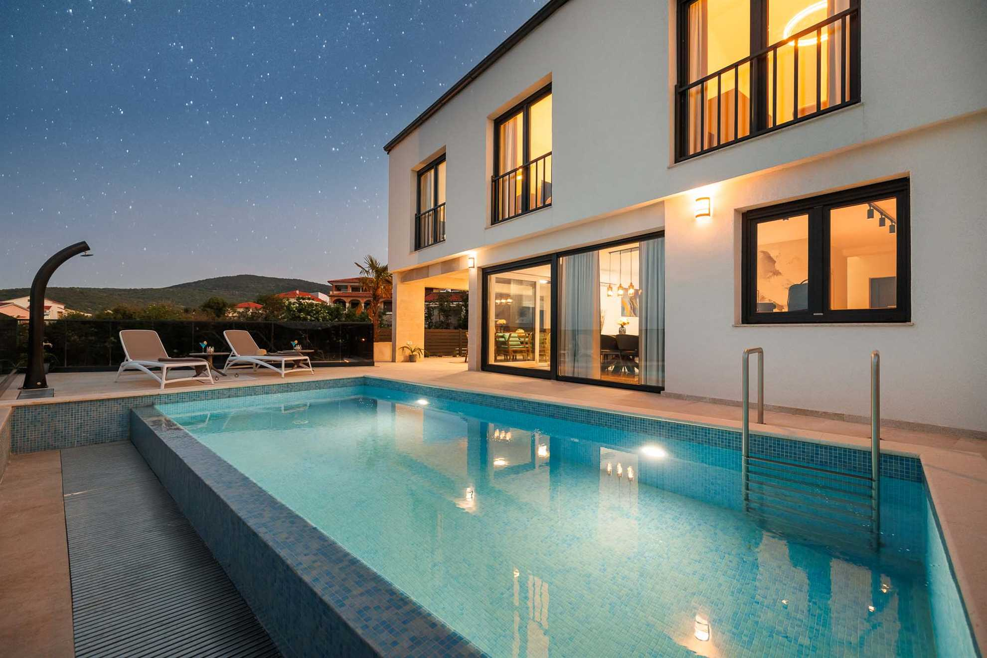 Illuminated luxury villa SUNSET I in Vrbnik with a heated pool, sun loungers, and mountain views. Nighttime ambiance.