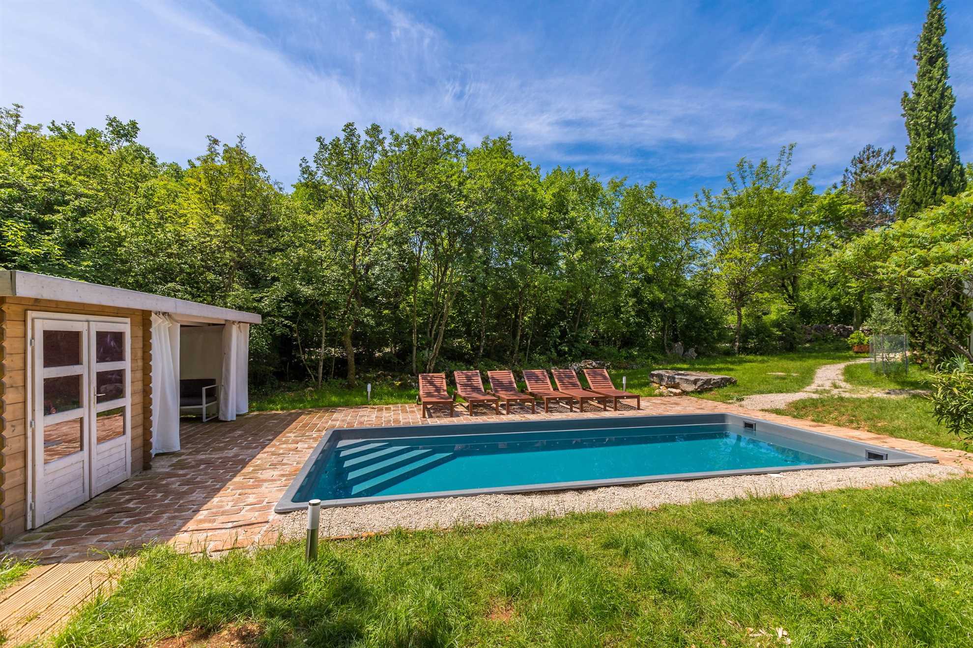 Swimming pool with sun loungers. Net to the pool is a small house. The pool is surrounded by trees.